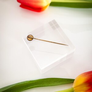 May include: A vintage gold-toned hat pin with a circular decorative head, resting on a clear acrylic block. The pin is set against a white background, with red and yellow tulips and green leaves in the background.