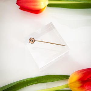 May include: A gold-toned lapel pin with a circular, patterned head featuring a central design, set on a clear acrylic block. The pin is surrounded by red and yellow tulips with green leaves, creating a vibrant contrast against the white background.