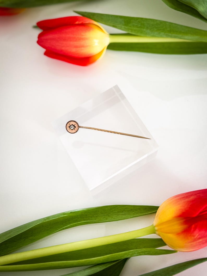 May include: A gold-toned lapel pin with a circular, patterned head featuring a central design, set on a clear acrylic block. The pin is surrounded by red and yellow tulips with green leaves, creating a vibrant contrast against the white background.