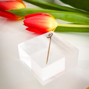 May include: A gold-toned stick pin with a circular, textured head featuring a small, central gemstone. The pin is displayed on a clear, rectangular block, with red and yellow tulips and green leaves in the background.