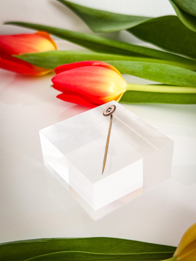 May include: A gold-toned stick pin with a circular, textured head featuring a small, central gemstone. The pin is displayed on a clear, rectangular block, with red and yellow tulips and green leaves in the background.