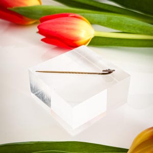 May include: A gold-colored hat pin with a decorative head rests on a clear acrylic block. The pin is set against a backdrop of red and yellow tulips with green leaves, creating a contrast of colors and textures.