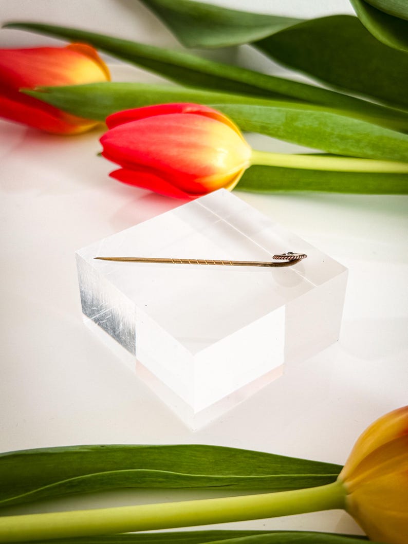 May include: A gold-colored hat pin with a decorative head rests on a clear acrylic block. The pin is set against a backdrop of red and yellow tulips with green leaves, creating a contrast of colors and textures.