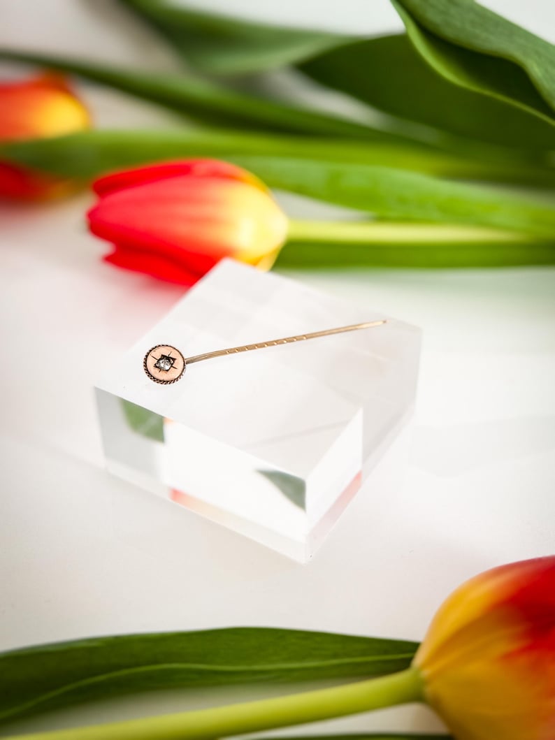 May include: A gold-toned lapel pin with a circular design featuring a small gemstone, displayed on a clear acrylic block. The pin is set against a backdrop of red and yellow tulips and green leaves, creating a contrast of textures and colors.
