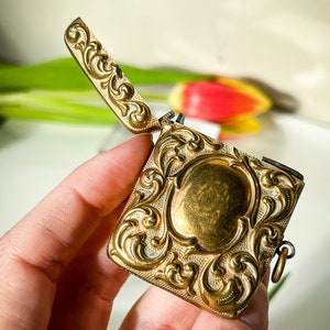 K&ouml;nnte beinhalten: Ein verziertes, goldfarbenes Medaillon mit einem dekorativen floralen Design. Das Medaillon ist ge&ouml;ffnet und hat eine kleine Kette daran.