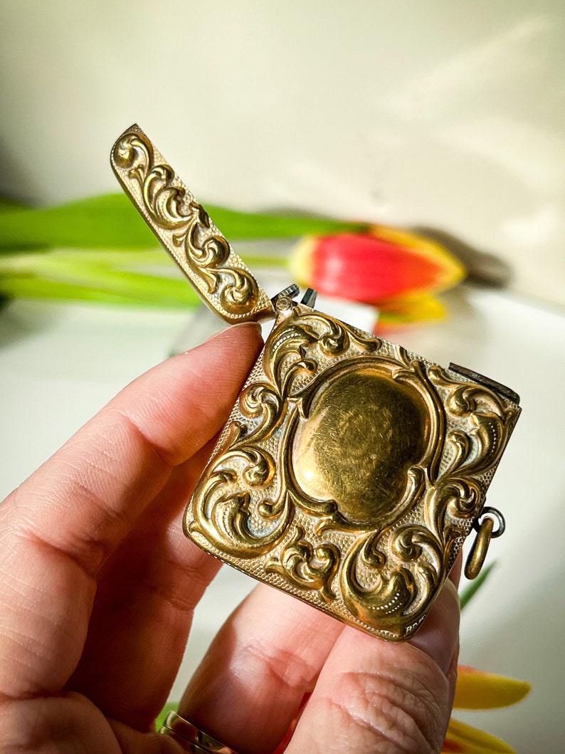 K&ouml;nnte beinhalten: Ein verziertes, goldfarbenes Medaillon mit einem dekorativen floralen Design. Das Medaillon ist ge&ouml;ffnet und hat eine kleine Kette daran.