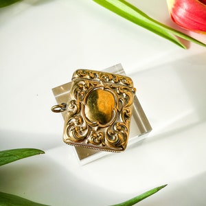 K&ouml;nnte beinhalten: Ein verziertes, goldfarbenes Streichholzschachtel mit einem dekorativen Blumendesign. Die Streichholzschachtel hat eine Schlaufe zum Anbringen an einer Kette.