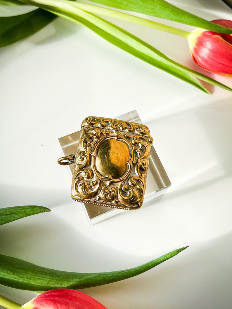 K&ouml;nnte beinhalten: Ein verziertes, goldfarbenes Streichholzschachtel mit einem dekorativen Blumendesign. Die Streichholzschachtel hat eine Schlaufe zum Anbringen an einer Kette.