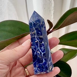 Puede incluir: Una punta de cristal de sodalita azul y blanca, también conocida como varita de cristal, se sostiene en una mano. El cristal tiene una punta puntiaguda y una base plana. El cristal es una piedra natural con un patrón de mármol azul y blanco.