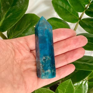 Puede incluir: Una punta de cristal azul y verde, también conocida como varita de cristal, se sostiene en una mano. El cristal es liso y pulido.