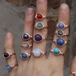 May include: A hand wearing a variety of rings with different colored gemstones. The rings are made of rose gold wire and feature a variety of stones, including amethyst, turquoise, carnelian, and lapis lazuli.