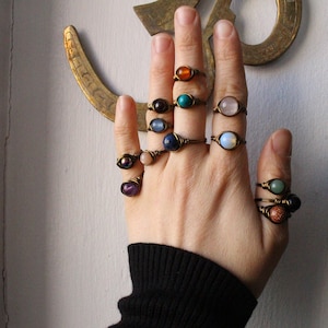 May include: A hand wearing a variety of rings with different colored gemstones. The rings are made of brass wire and feature a variety of stones, including amethyst, turquoise, and carnelian.
