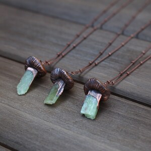 May include: Three copper necklaces with green crystal pendants shaped like mushrooms. The necklaces are on a wooden surface.