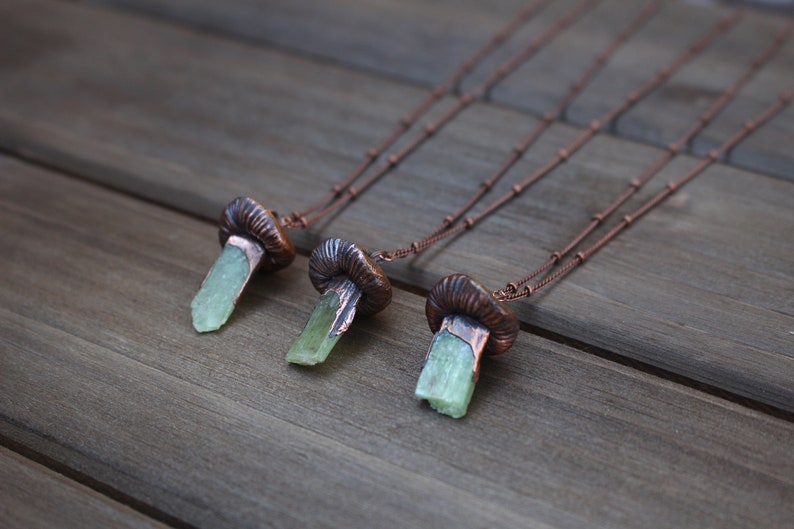 May include: Three copper necklaces with green crystal pendants shaped like mushrooms. The necklaces are on a wooden surface.