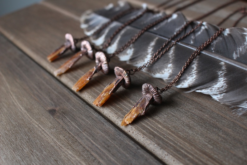 May include: Five necklaces with copper mushroom pendants and orange crystal points. The necklaces are displayed on a wooden surface with a feather.