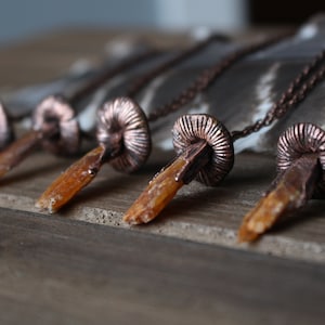 May include: Five copper and crystal necklaces with a mushroom-shaped pendant. Each necklace features a different crystal point, all with a similar orange-brown color.
