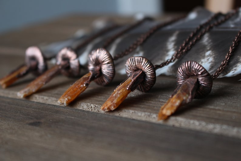 May include: Five copper and crystal necklaces with a mushroom-shaped pendant. Each necklace features a different crystal point, all with a similar orange-brown color.