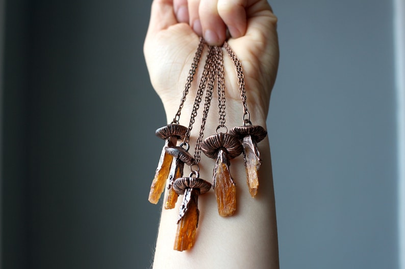 May include: A set of five copper necklaces with orange crystal pendants. Each pendant is shaped like a mushroom with a copper cap and a long, thin crystal stem.