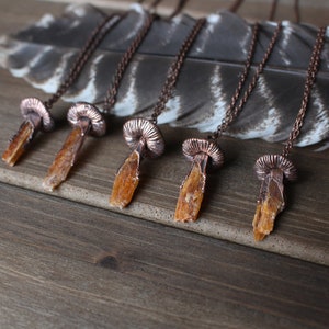 May include: Five copper necklaces with orange crystal pendants shaped like mushrooms. The necklaces are displayed on a wooden surface with a feather.