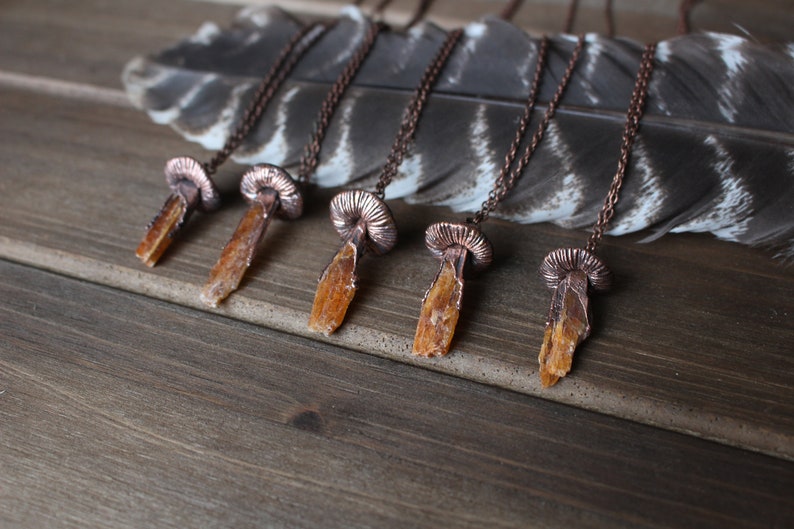 May include: Five copper necklaces with orange crystal pendants shaped like mushrooms. The necklaces are displayed on a wooden surface with a feather.