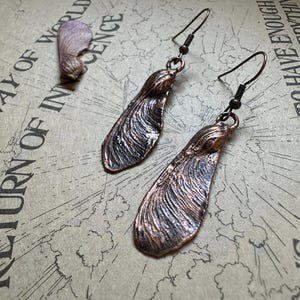 May include: Copper-colored earrings in the shape of maple tree seeds, with detailed veining. The earrings hang from dark metal ear wires. A single seed and a vintage map background complete the image.