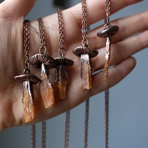 May include: Five copper necklaces with mushroom pendants and orange crystal charms. The necklaces are displayed on a hand.