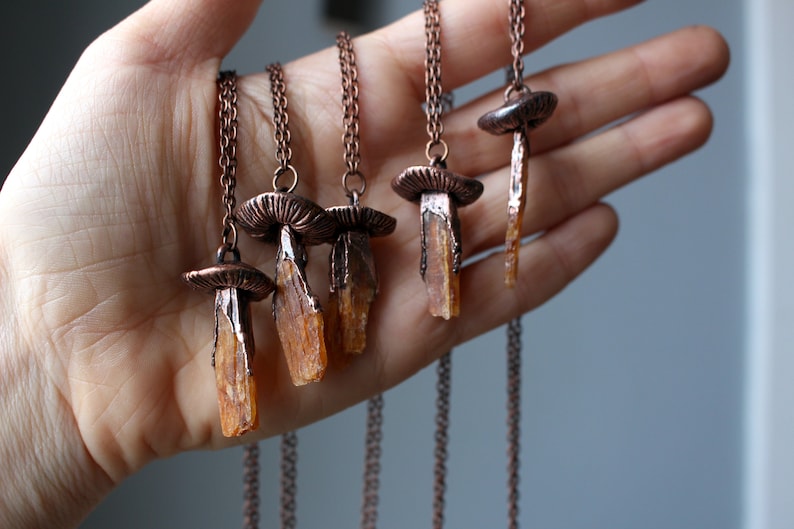 May include: Five copper necklaces with mushroom pendants and orange crystal charms. The necklaces are displayed on a hand.
