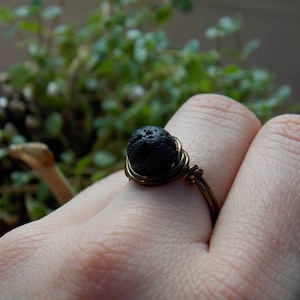 May include: A black lava rock ring with a brass wire wrap. The ring is on a person's finger.