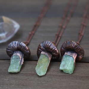 May include: Three copper mushroom necklaces with green crystal pendants. The necklaces are on a wooden surface.