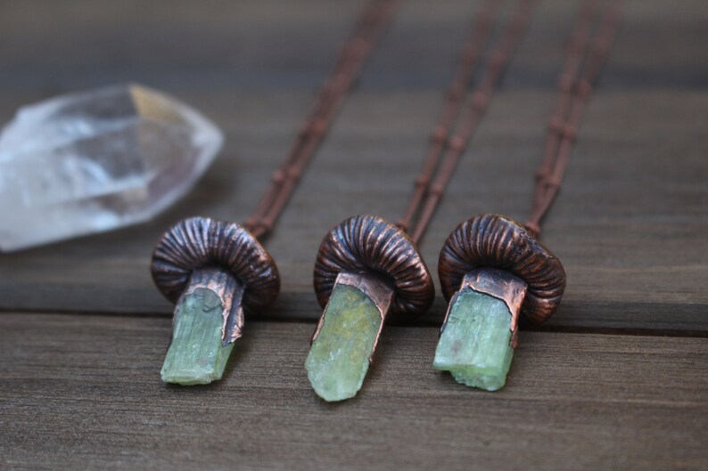 May include: Three copper mushroom necklaces with green crystal pendants. The necklaces are on a wooden surface.