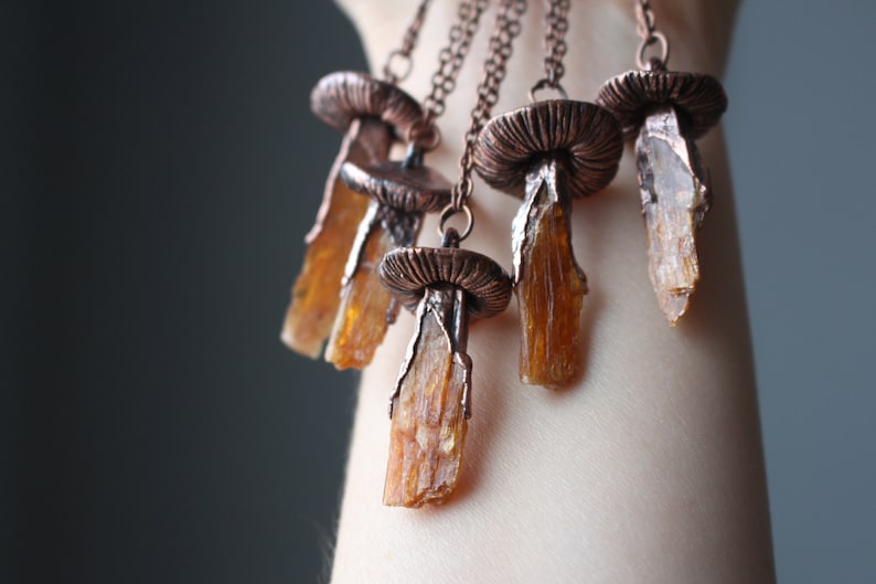 May include: A necklace with five copper mushroom pendants, each with a clear amber crystal hanging from the bottom. The necklace is on a person's arm.