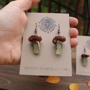 May include: Copper mushroom earrings with green crystal accents. The earrings are on a card with the text "Gratefully Dreaming Etsy.com".
