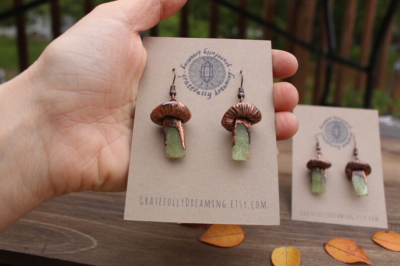 May include: Copper mushroom earrings with green crystal accents. The earrings are on a card with the text "Gratefully Dreaming Etsy.com".