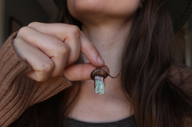 May include: A copper pendant necklace with a light green crystal. The crystal is a rectangular shape and is set in a copper frame. The necklace is on a chain.