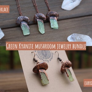 May include: A set of green kyanite mushroom jewelry, including a necklace and earrings. The necklace features three copper mushroom pendants with green kyanite stones. The earrings are copper mushroom dangles with green kyanite stones. The jewelry is displayed on a wooden surface with clear quartz crystals.