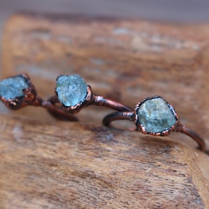 May include: Three copper-toned rings, each featuring a rough-cut, light blue gemstone. The rings are displayed on a piece of weathered wood, showcasing the handcrafted jewelry's natural aesthetic. The gemstones have a unique, organic shape.