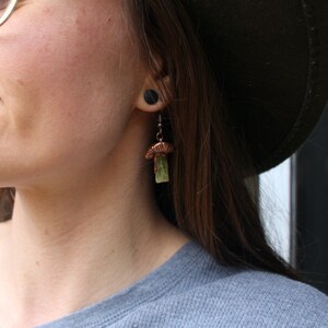 May include: A pair of copper mushroom earrings with green stone accents. The earrings are hanging from the earlobe of a person with brown hair.