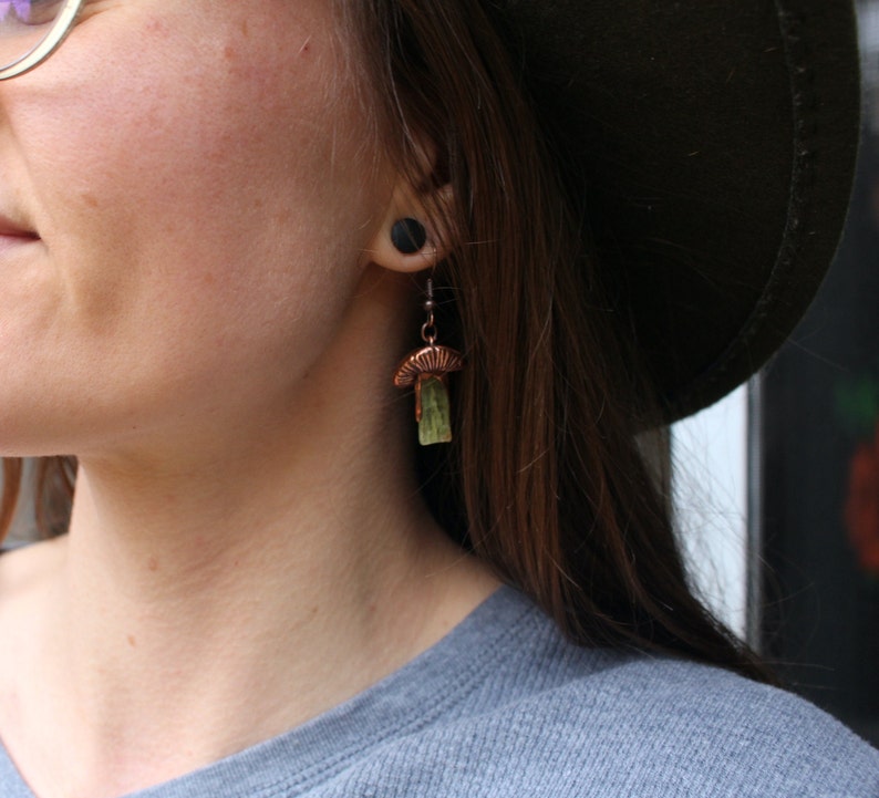 May include: A pair of copper mushroom earrings with green stone accents. The earrings are hanging from the earlobe of a person with brown hair.