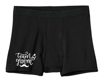 May include: Black boxer shorts with the text "Team Groom" in white lettering with a ring and a mustache.