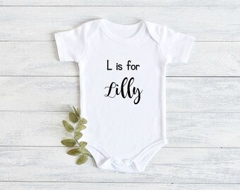 May include: White baby onesie with black text that reads "L is for Lilly".