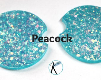 May include: Two turquoise blue round coasters with a cut-out for a cup. The coasters are covered in iridescent glitter and the text "Peacock" is printed in black in the center of the image.