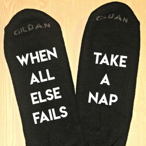 K&ouml;nnte beinhalten: Paar schwarze Socken mit wei&szlig;em Text. Eine Socke liest "WHEN ALL ELSE FAILS" und die andere "TAKE A NAP". Der Markenname "GILDAN" ist oben auf jeder Socke aufgedruckt.