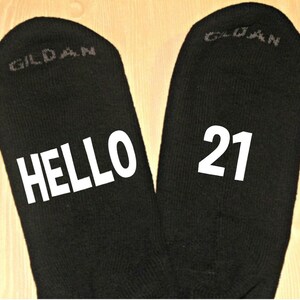 May include: Black socks with the text "HELLO 21" in white. The socks are made of a dark material and are displayed on a light wood surface. The brand name "GILDAN" is printed on the socks.