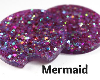 May include: Two round purple glitter coasters with a cutout for a cup. The coasters have a iridescent shimmer and the text "Mermaid" is written below them.