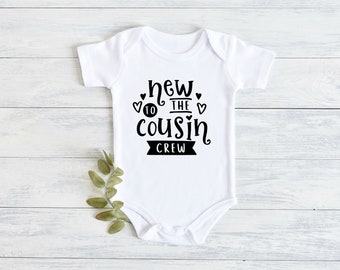 May include: White baby onesie with black text that reads "new to the cousin crew" with two hearts and a number 10.