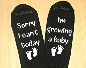 May include: A pair of black socks with white text. One sock says "Sorry I can't today" with a pair of white footprints. The other sock says "I'm growing a baby" with a pair of white footprints.