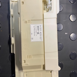 May include: A beige and white electronic component with the text "9 001 409 627" and "WKKT" printed on a label. Additional text includes "07.03.23 06:43 27" and "Typ PW3(120)V AC". The component is rectangular with various protruding parts.