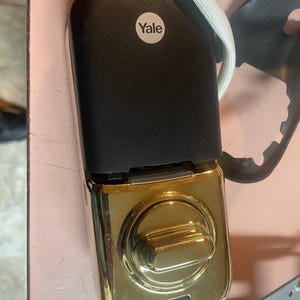 May include: A smart lock with a black top and a gold-colored bottom. The lock has a round Yale logo sticker and a white cord attached. The lock is designed for home security.