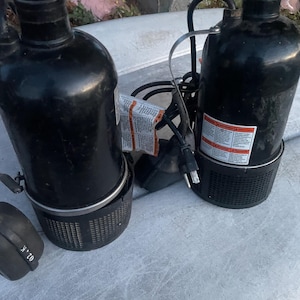 May include: Two black submersible water pumps with a cylindrical design. Each pump has a black mesh base and a power cord. One pump has a warning label attached. The pumps are on a concrete surface.
