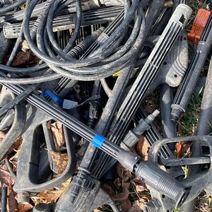 May include: A pile of black pressure washer parts, including hoses, spray wands, and handles. The components are scattered, with some showing signs of wear. A blue band is visible on one of the spray wands.